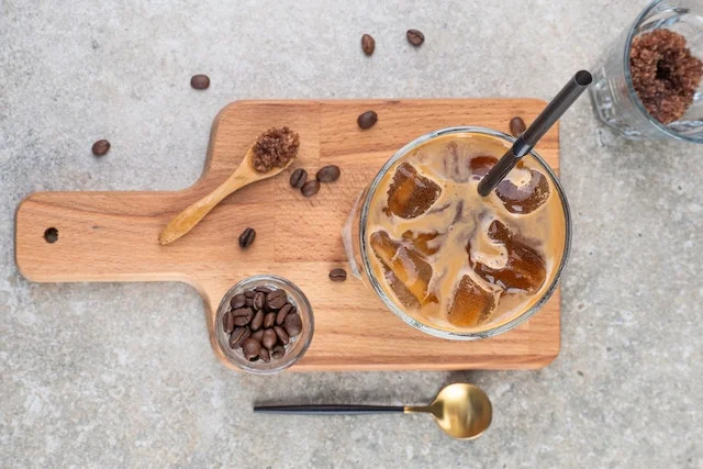 Glass of iced coffee on a chopping board with coffee beans scattered around it.