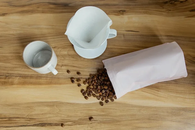 Coffee beans falling out of a paper bag on a table next to a milk jug and coffee cup, illustrating sustainable coffee packaging.